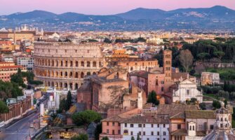 Skyline of Rome, Italy at sunset, with historic buildings including the Colosseum in the foreground, and mountains in the background