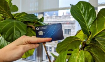 The Chase Sapphire Preferred Card held up before a leafy plant with buildings seen through the window in the background