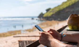 Close up on a man's hands holding a cell phone with a coconut and beach in the background