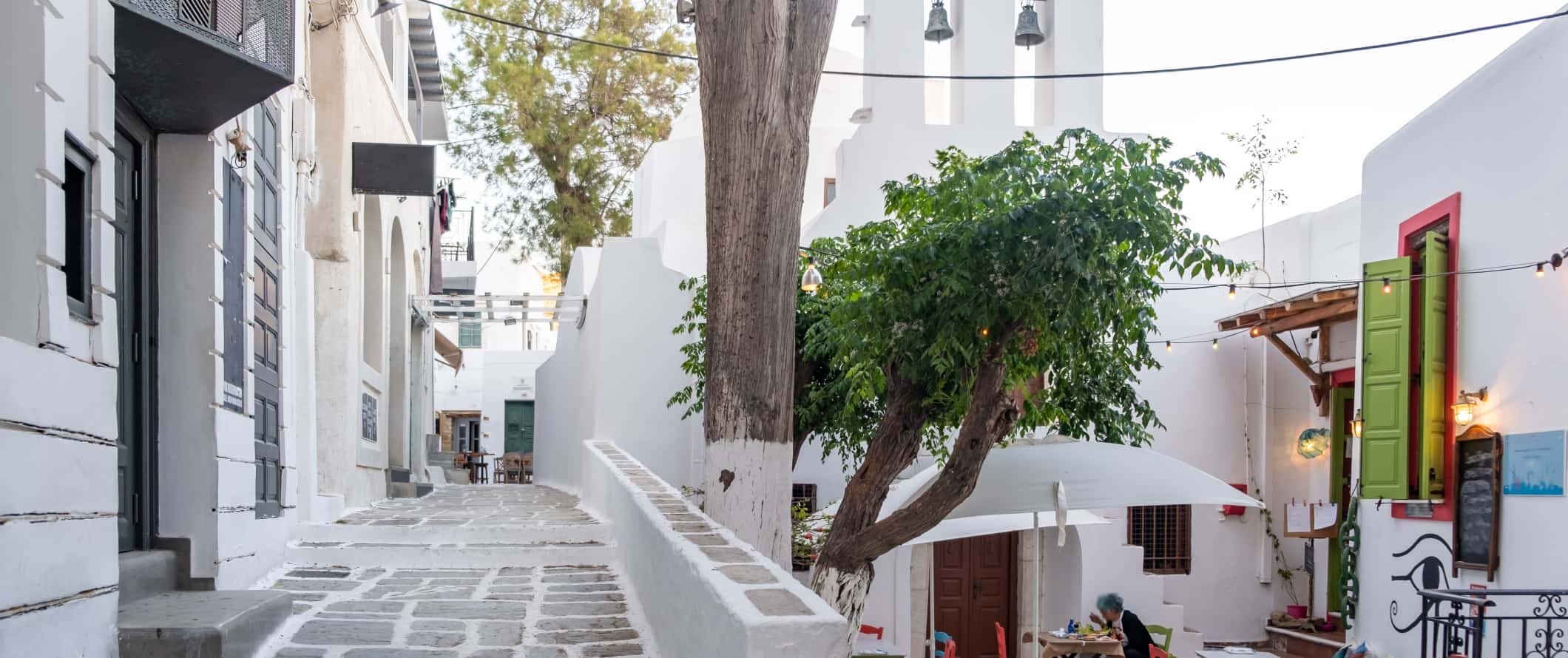 View of white-washed buildings, a street lined with flagstones, and outdoor seating at a cafe with brightly colored tables and chairs in Ios, Greece