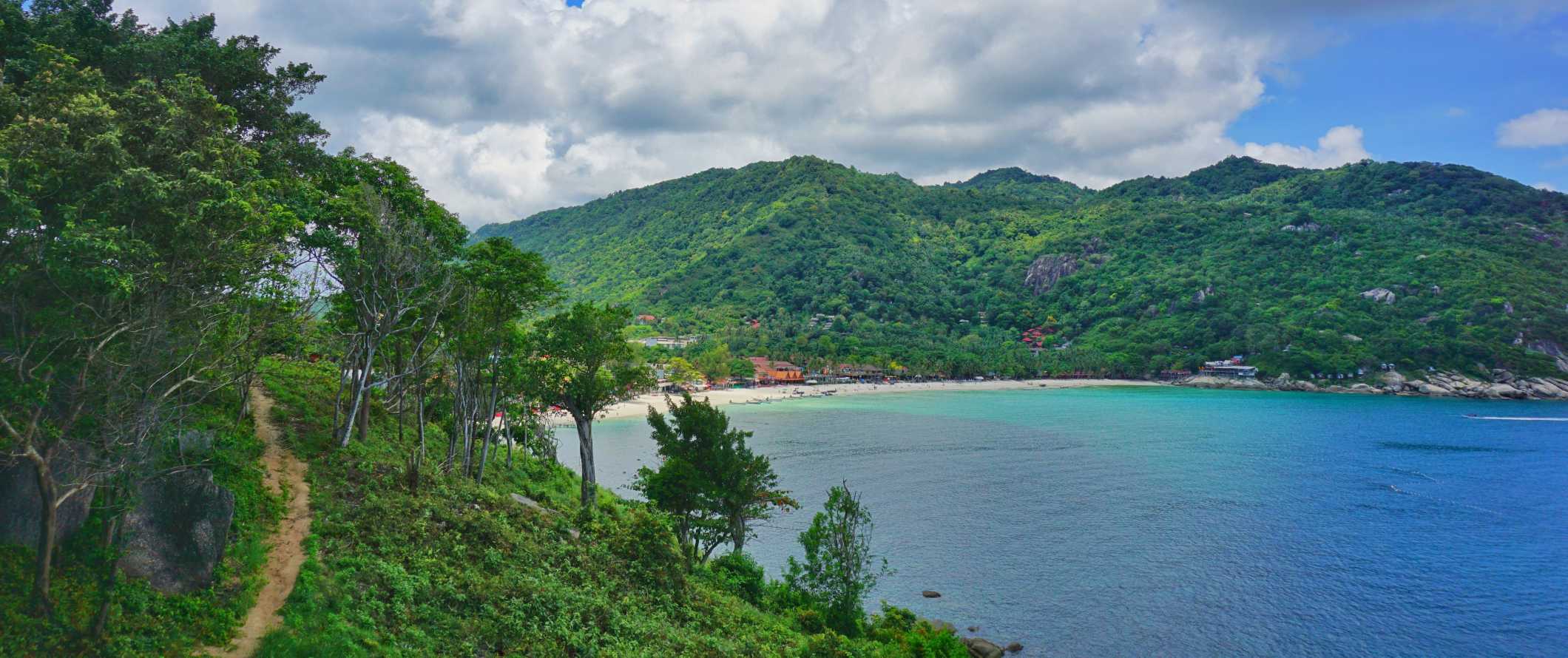 A hiking path going through the lush forest with a turquoise bay in the background on the island of Ko Pha Ngan, Thailand at sunset over the ocean