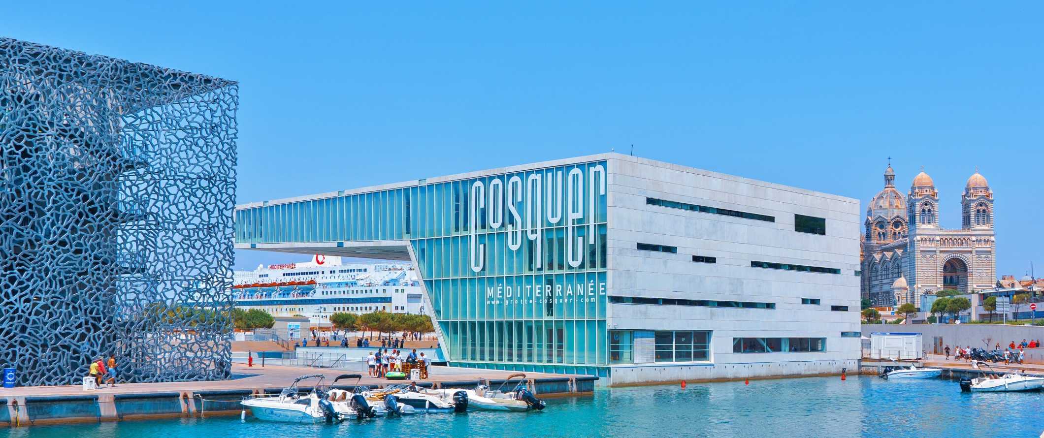 Motorboats docked in front of futuristic buildings and a large cruise ship looming in the background in Marseille, France