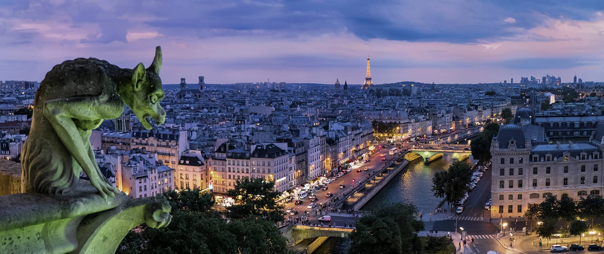 A gargoyle in the foreground on an old building in Paris during a pink sunset over the city’s skyline