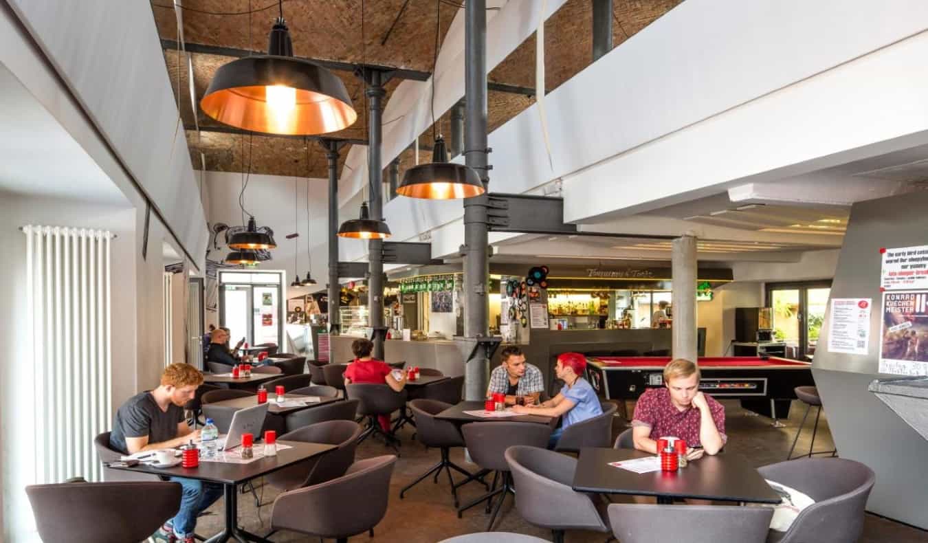 Common area with high ceilings and people sitting at tables working on laptops and talking at Pfefferbett Hostel, Berlin