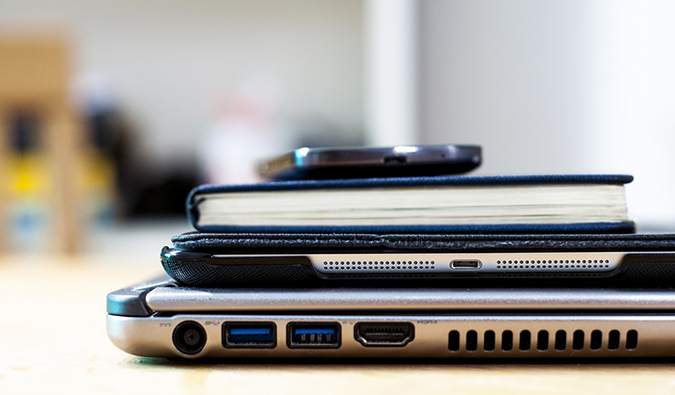 A tablet piled on a desk with a laptop, book, and smartphone